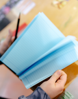 Hands flipping through a blue paper socolo A5 notebook at a wooden desk.